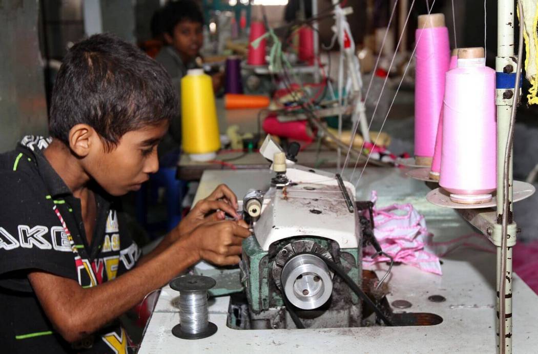 A child working at the sewing machine in a garment factory. Another child worker is seen in the background.